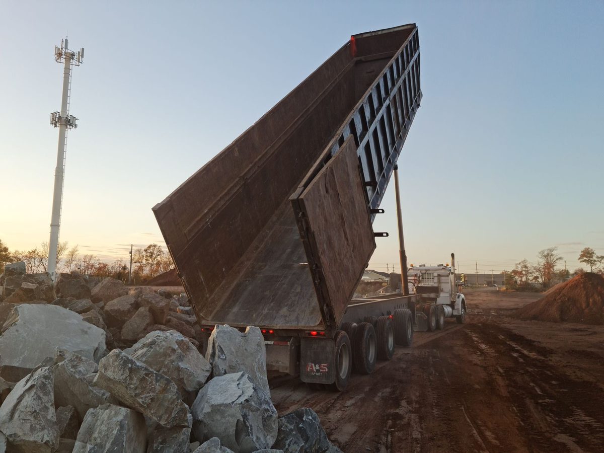 A dump truck with its bed raised, tipping a load of rock at dusk